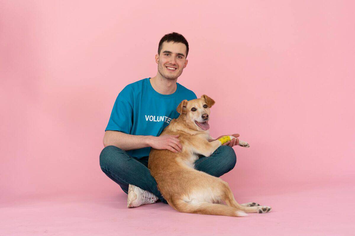 Jeune homme souriant en chemise de bénévole bleue assis avec un chien heureux sur fond rose.